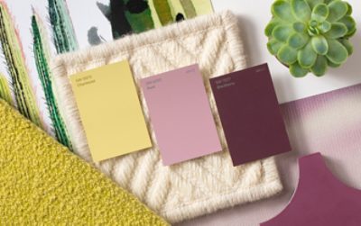 Yellow, pink, and berry colored paint chips laid out on a cream woven material, near other fabrics and a plant.