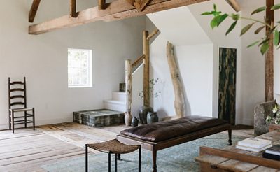 Interior of residential living space with white walls, natural wood beams and banisters in corner stairwell with collection of decorative ceramic vessels on the floor and leather-topped wood bench in foreground.