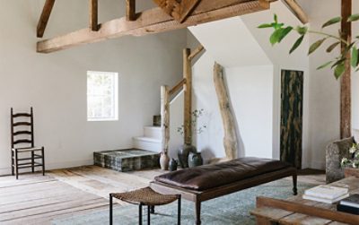 Interior of residential living space with white walls, natural wood beams and banisters in corner stairwell with collection of decorative ceramic vessels on the floor and leather-topped wood bench in foreground.