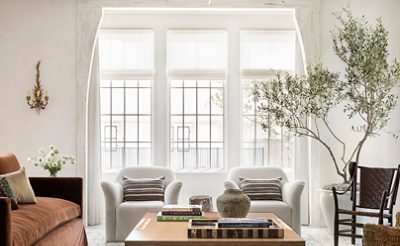 Elegant living room with a brown sofa, white armchairs, large windows, coffee table with books, and a stone fireplace with mirrors and a backdrop of walls painted Greek Villa by Sherwin-Williams.