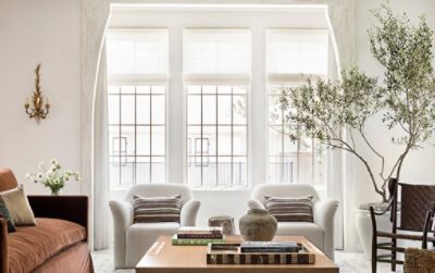 Elegant living room with a brown sofa, white armchairs, large windows, coffee table with books, and a stone fireplace with mirrors and a backdrop of walls painted Greek Villa by Sherwin-Williams.