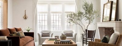 Elegant living room with a brown sofa, white armchairs, large windows, coffee table with books, and a stone fireplace with mirrors and a backdrop of walls painted Greek Villa by Sherwin-Williams.