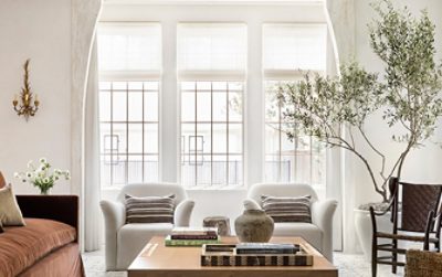 Elegant living room with a brown sofa, white armchairs, large windows, coffee table with books, and a stone fireplace with mirrors and a backdrop of walls painted Greek Villa by Sherwin-Williams.