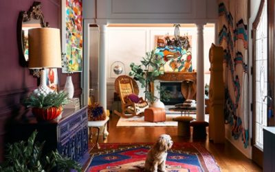 Colorful interior with eclectic decor, including art, plants, a wooden rocking chair, and a dog on a patterned rug.