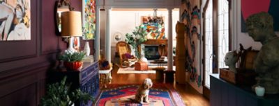 Colorful interior with eclectic decor, including art, plants, a wooden rocking chair, and a dog on a patterned rug.