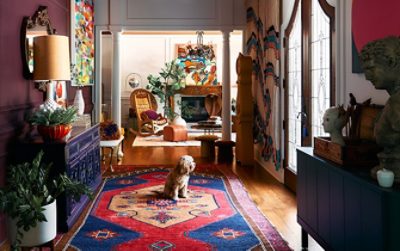 Colorful interior with eclectic decor, including art, plants, a wooden rocking chair, and a dog on a patterned rug.