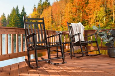 Two black wooden rocking chairs on a stained deck on a lake with colorful fall foliage behind.