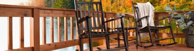 Two black wooden rocking chairs on a stained deck on a lake with colorful fall foliage behind.