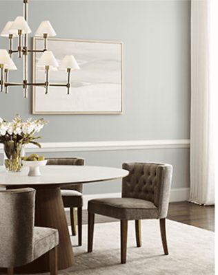 Dining room with large round table and cushioned chairs, light fixture and painting.