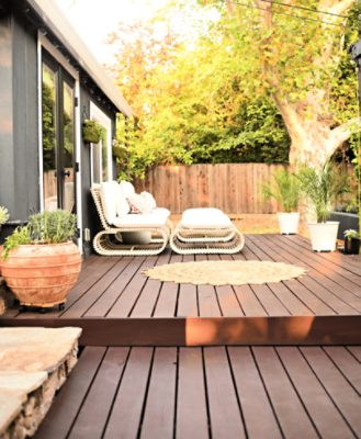 A deck stained a dark red-brown with potted plants, a circular outdoor rug and white modern furniture.