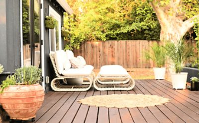 A deck stained a dark red-brown with potted plants, a circular outdoor rug and white modern furniture.