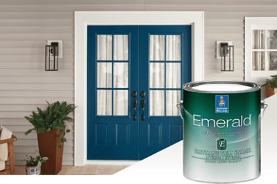A can of exterior Emerald paint set in front of double front doors painted blue with a potted fern to the left side and an outdoor chair.