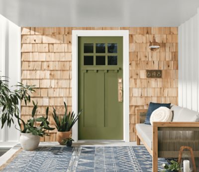 A porch with a bench and some plants with a green door and light wood siding.