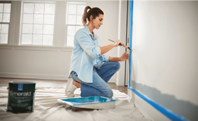 A person kneeling while painting a white wall next to a roller trey and can of paint.