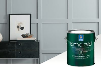 A can of interior Emerald paint set in front of a gray painted wall with a small table with an abstract print and succulents on top.