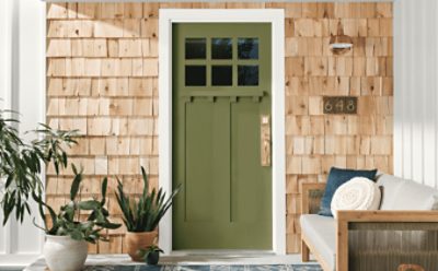 Front door painted green with wooden siding surrounding, couch and plants to fill the porch.
