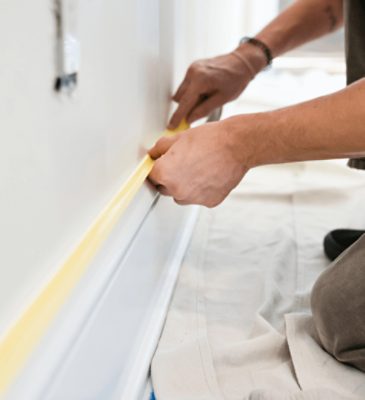 A person kneeling on a drop cloth applying masking tape to a white baseboard.