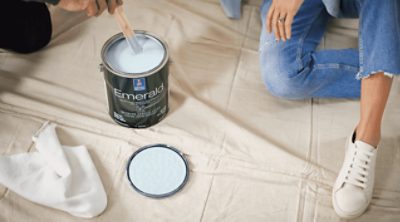A person kneeling on a cream drop cloth next to a can of Sherwin-Williams Emerald paint.
