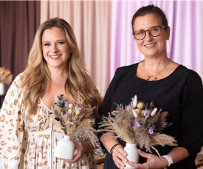 A photo of two people each holding a ceramic vase with dried wheat and flowers.