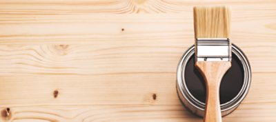 Paintbrush lying across an open can of wood stain on a light brown wooden floor, viewed from above.