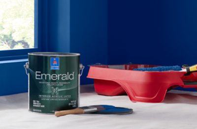 A can of Emerald Interior Acrylic Latex paint in front of a freshly painted blue wall and a red paint tray.