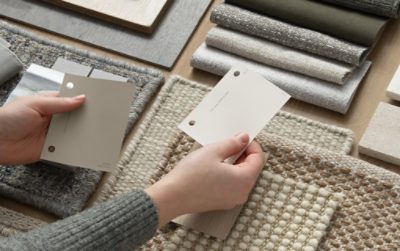 Fabric and textile swatches in a neutral color scheme and hands holding sample color chips in Sherwin-Williams colors Quarry Stone and Drift of Mist.