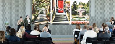 A group of people seated facing a projected image of three artworks displayed on a screen, representing the Roots, Soul, and Flair palettes of the Colors of Savannah project, with presenters standing nearby.