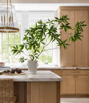 A bright kitchen with marble island, potted plant, wicker stools, wooden cabinets, and large windows. 