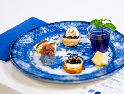A blue patterned plate featuring small bites and a plastic cup of tea with a mint sprig and lemon wedge. 