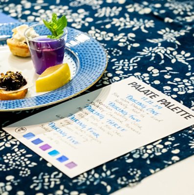 A plate with various appetizers inspired by the color blue and a menu card on a blue patterned tablecloth. 