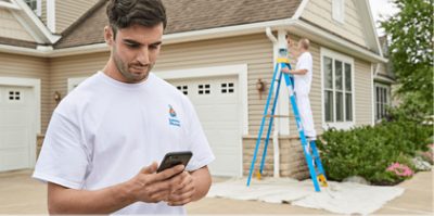A contractor looking at his smart phone in a driveway of a house being painted by another contractor.