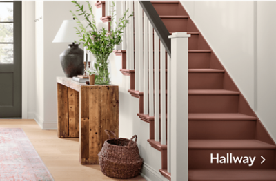 A hallway with white walls and natural accents next to a red-brown painted staircase.