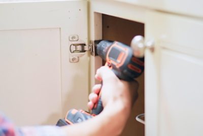 A person using a drill to remove hinges from a cabinet door.