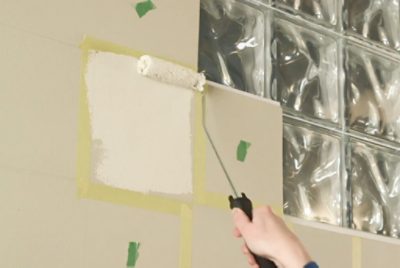 Hand painting a white square on a beige wall using a roller, with masking tape and a glass block window nearby.