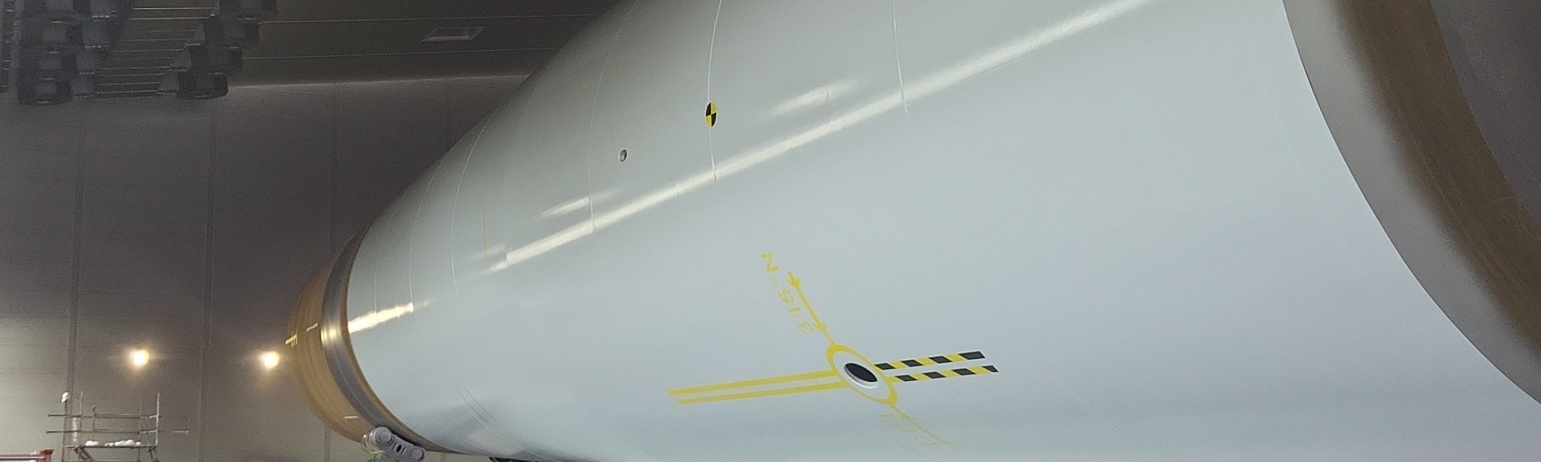 Large cylindrical wind turbine tower section inside an industrial coating facility, with a worker in high‑visibility safety gear standing nearby for scale.