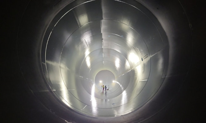 View inside a large cylindrical wind turbine tower section, showing a reflective metal interior with two workers standing at the far end for scale.