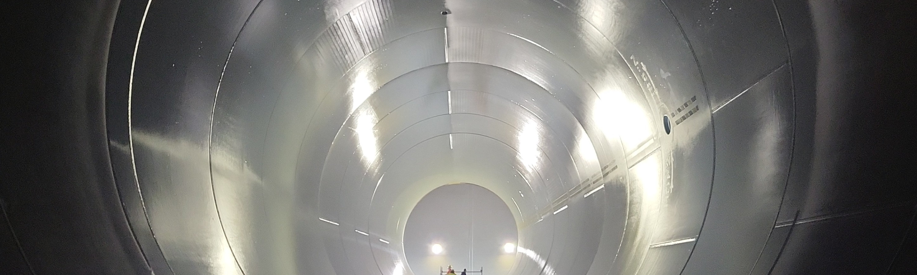 View inside a large cylindrical wind turbine tower section, showing a reflective metal interior with two workers standing at the far end for scale.
