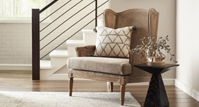 Cream walls, wooden floors with rug and tan chair with pillow next to a side table with flowers.