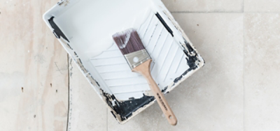A paint brush in a used paint tray filled with white paint on the floor.