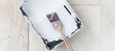 A paint tray on tile floor filled with white paint and a used paint brush on top.