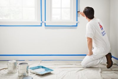 A paint pro painting alongside a window covered with blue painter's tape.