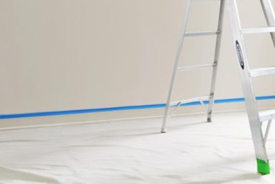 A ladder on top of a drop cloth in front of a wall with painters tape on the trim.