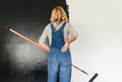 A person holding a roller on a long pole in front of a partially painted chalkboard wall.