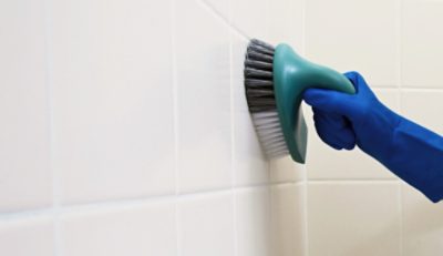 A person wearing a rubber glove while cleaning bathroom tile with a scrub brush.