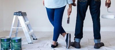 Two painters standing in front of a wall holding a paint brush and a paint roller.  Next to them is a ladder and two cans of Sherwin-Williams Emerald Paint.