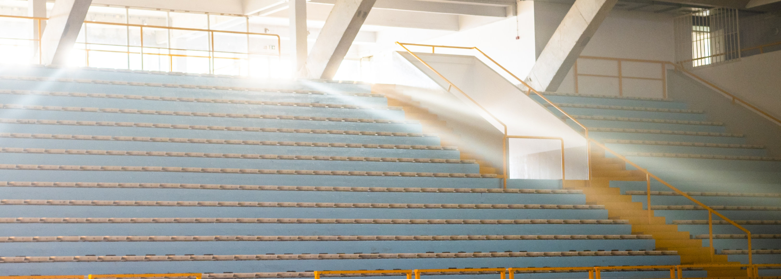 Indoor sports arena with blue seats, wooden floor, and sunlight streaming through windows.