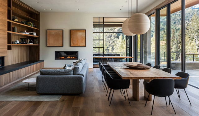 Modern open-plan living and dining area with large windows, grey sectional sofa, wooden table, and pendant lights. Sayerlack