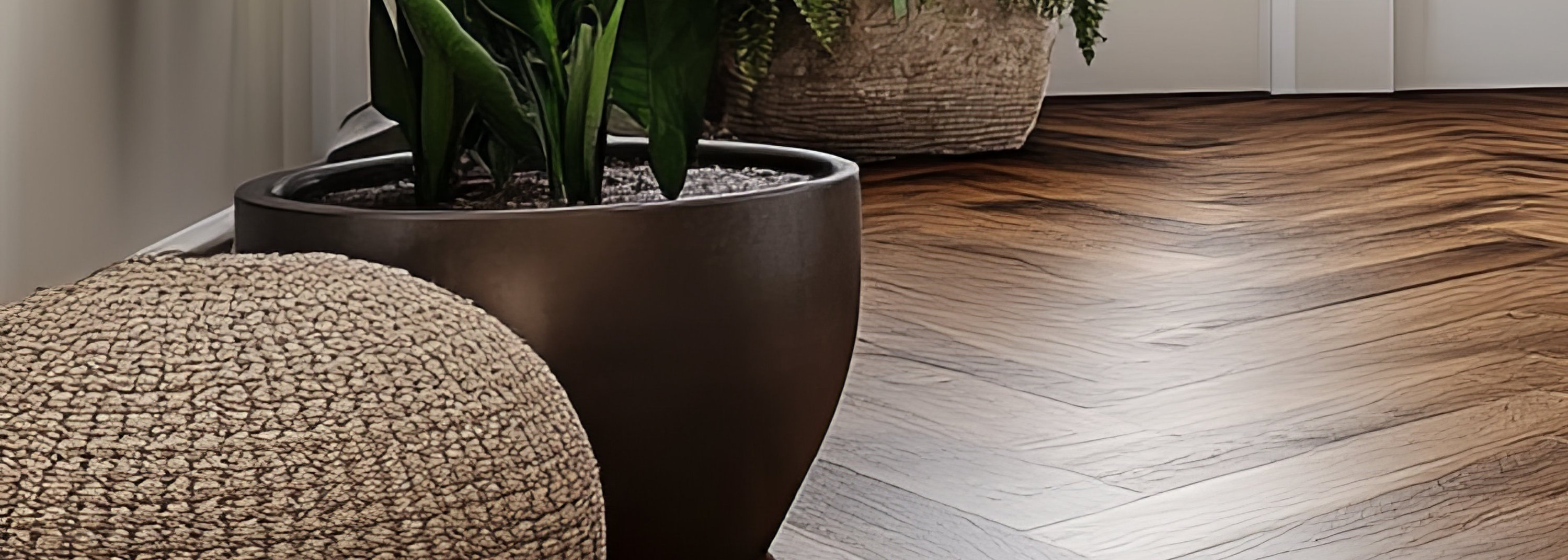 Cozy living room with herringbone wooden flooring, white sofa with cushions, potted plant, woven pouf, and natural light streaming through windows.