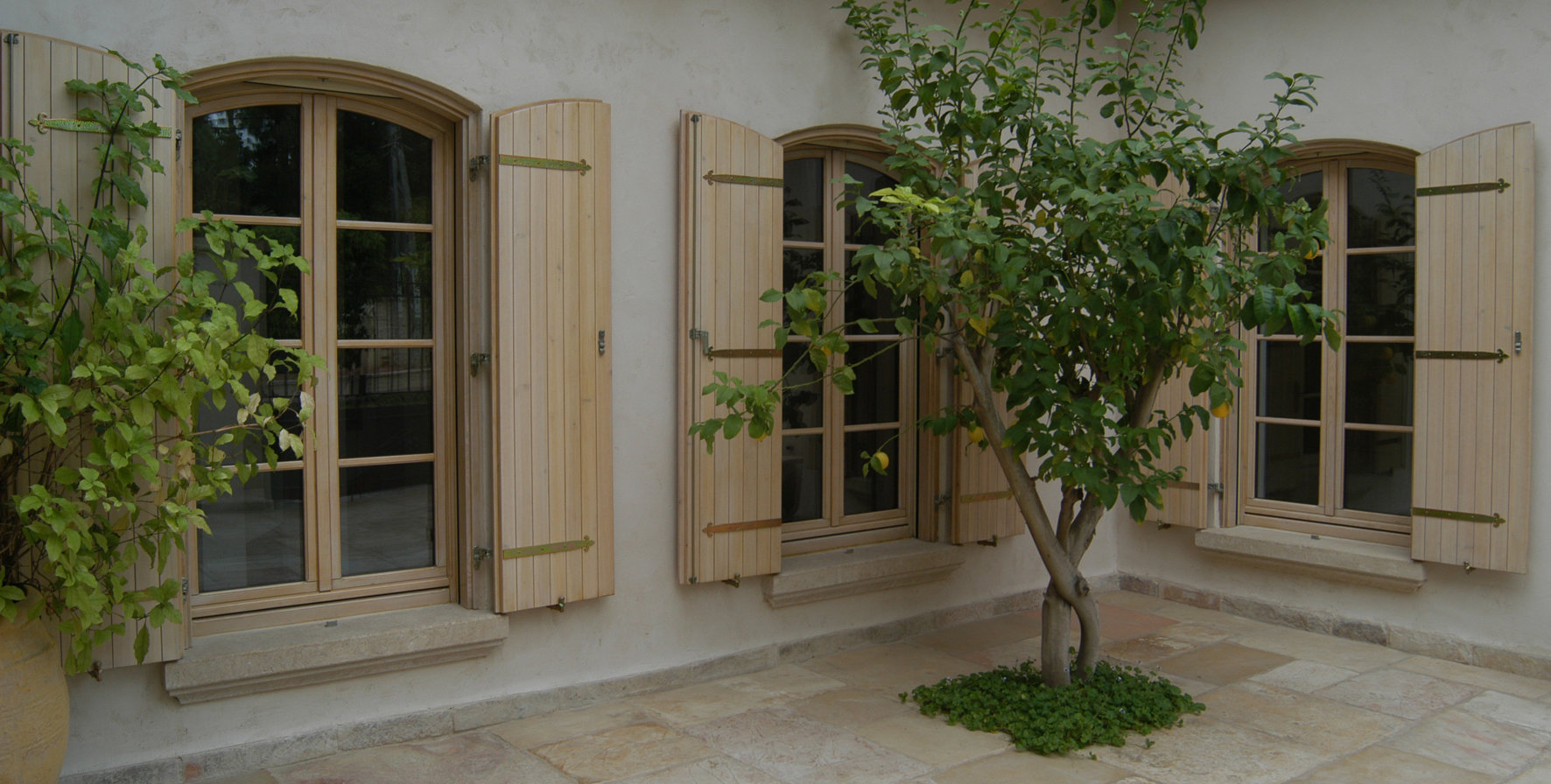 Courtyard with arched windows, open wooden shutters, a small tree, and potted plants on stone pavement.
