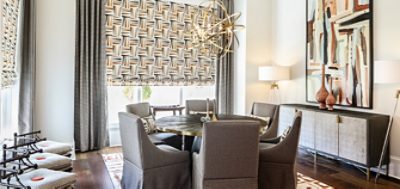 Boldly designed dining room with round table encircled by six upholstered dining chairs, patterned window coverings and modern gold orb chandelier.